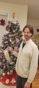 Tommy age 46 holds his batman socks from kindergarten in front of Christmas tree. He looks like he is tolerating his mom's memory. 